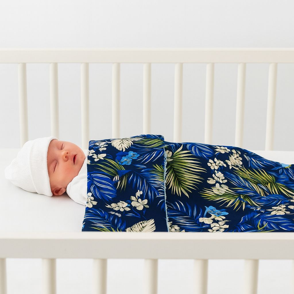 Babydecke tropisch blau – Babypuppe im weißen Strampler und Mütze liegt im Babybett, bedeckt mit weicher Baumwolldecke mit Blumen- und Blättermuster
