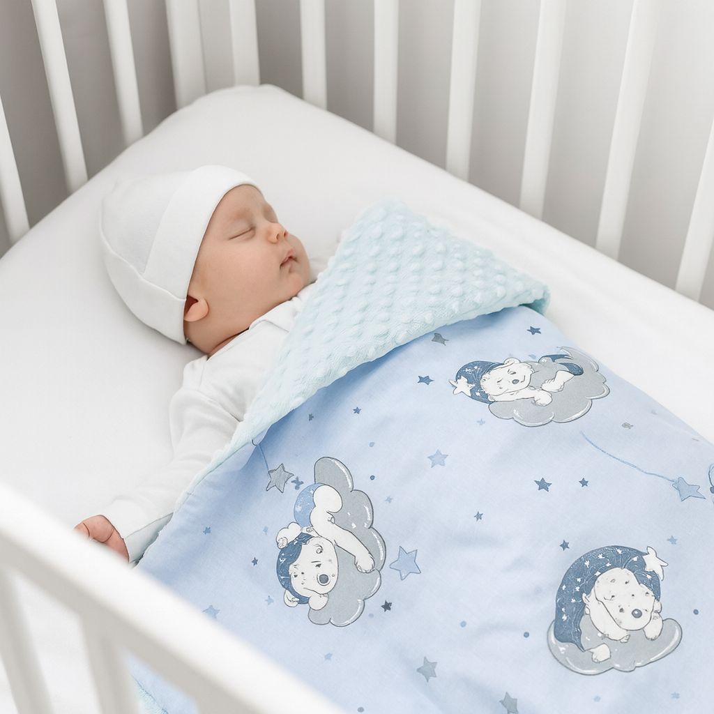 Schlafendes Neugeborenes liegt in einem weißen Babybett, trägt weiße Kleidung und Mütze, bedeckt mit einer hellblauen Babydecke mit Bärenmotiv